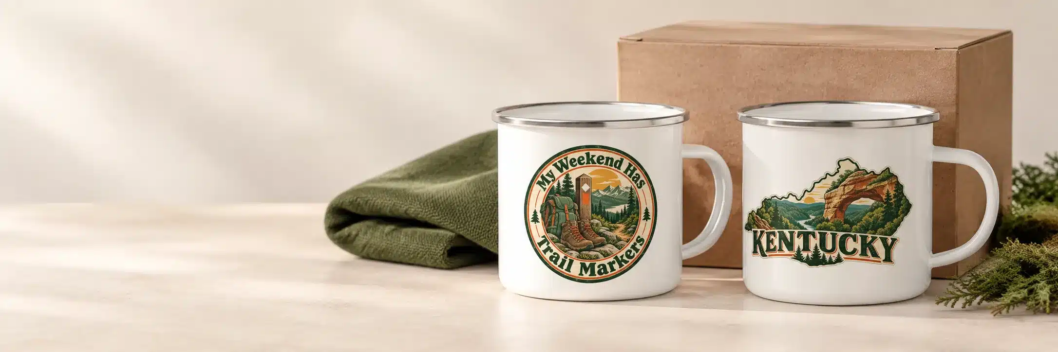 Two enamel camp mugs, one reading My Weekend Has Trail Markers and one with a Kentucky design, next to a kraft gift box and a green beanie